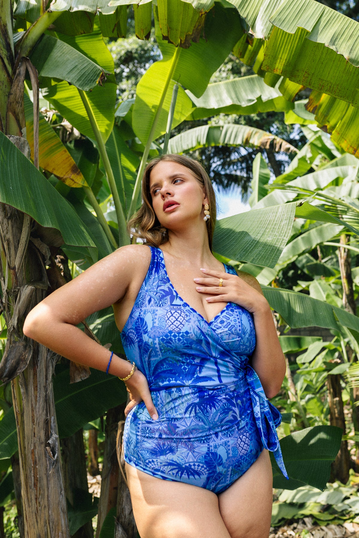 Woman standing in front of tropical palms wearing an ultramarine blue wrapsuit with vibrant lighter blue shades highlighting the Italian motifs inspired print.